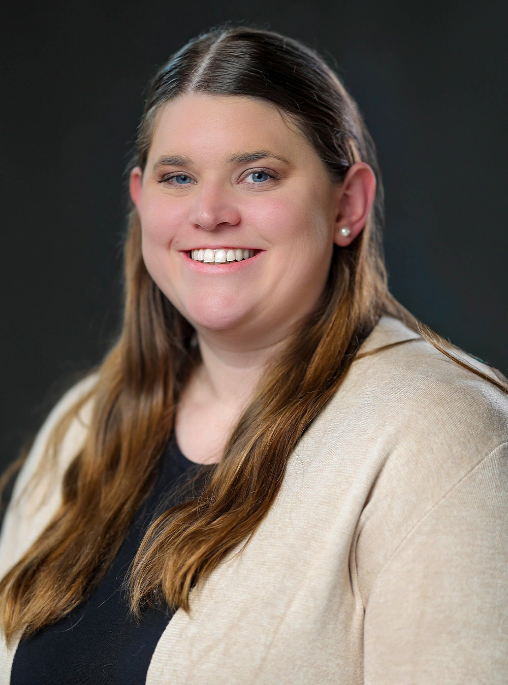 Portrait of a smiling woman with long brown hair wearing a beige jacket against a dark background