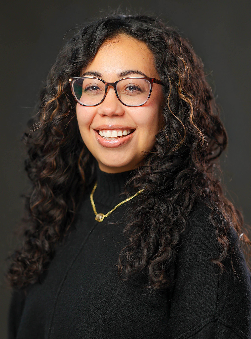 Portrait of a smiling woman with long brown hair wearing a black top against a dark background
