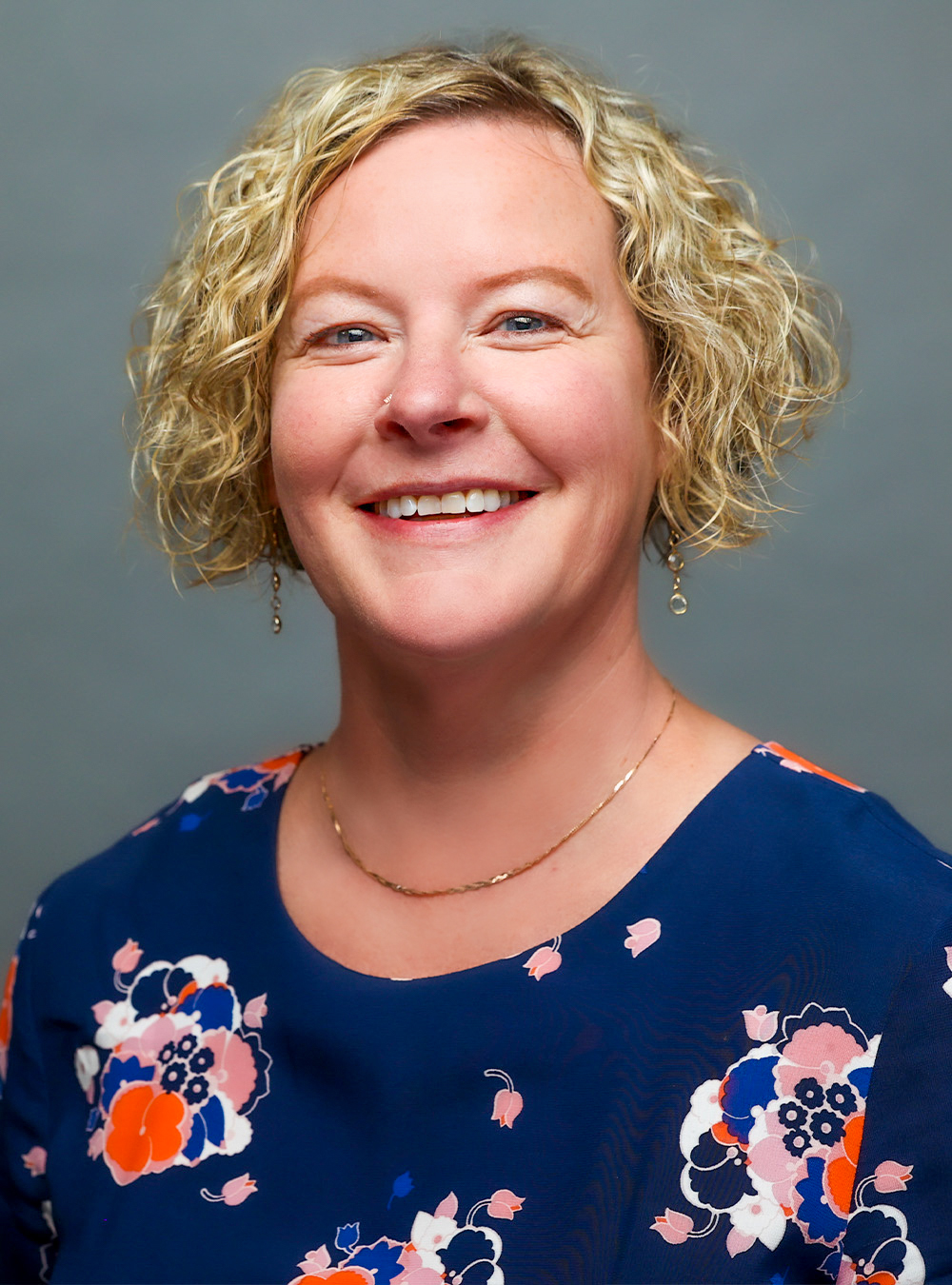 Portrait of a smiling woman with short blonde curly hair wearing a floral top against a neutral background
