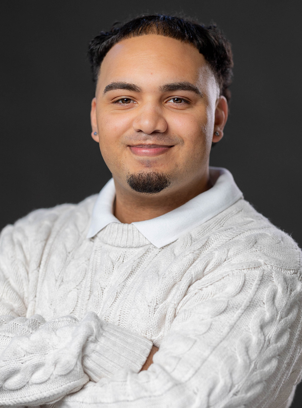 Portrait of a smiling man with short black hair wearing a neutral sweater against a dark background