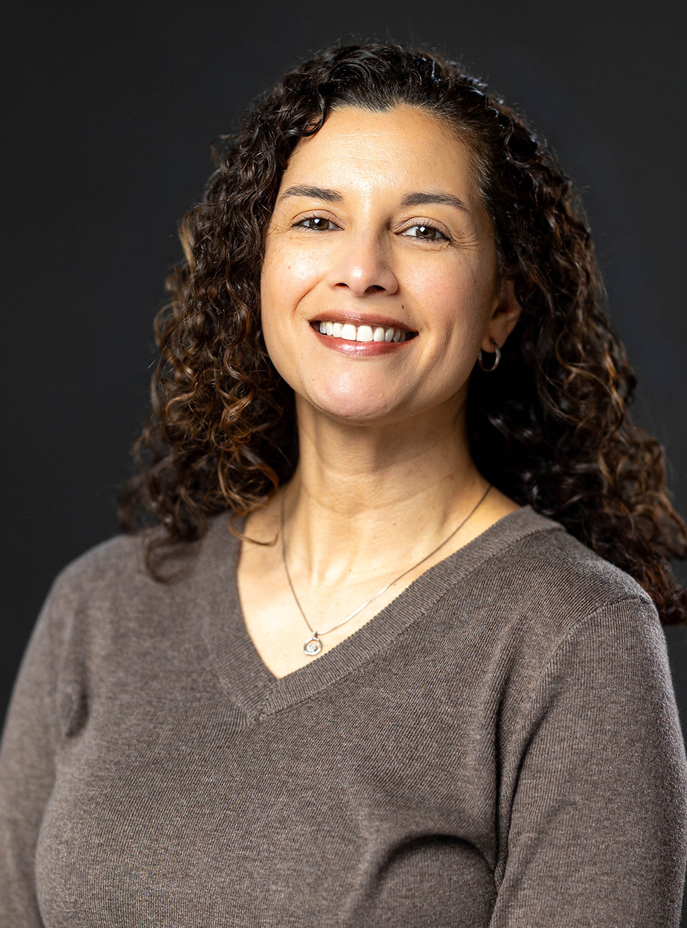 Portrait of a smiling woman with sholder-length dark hair wearing a brown top, smiling against a dark background
