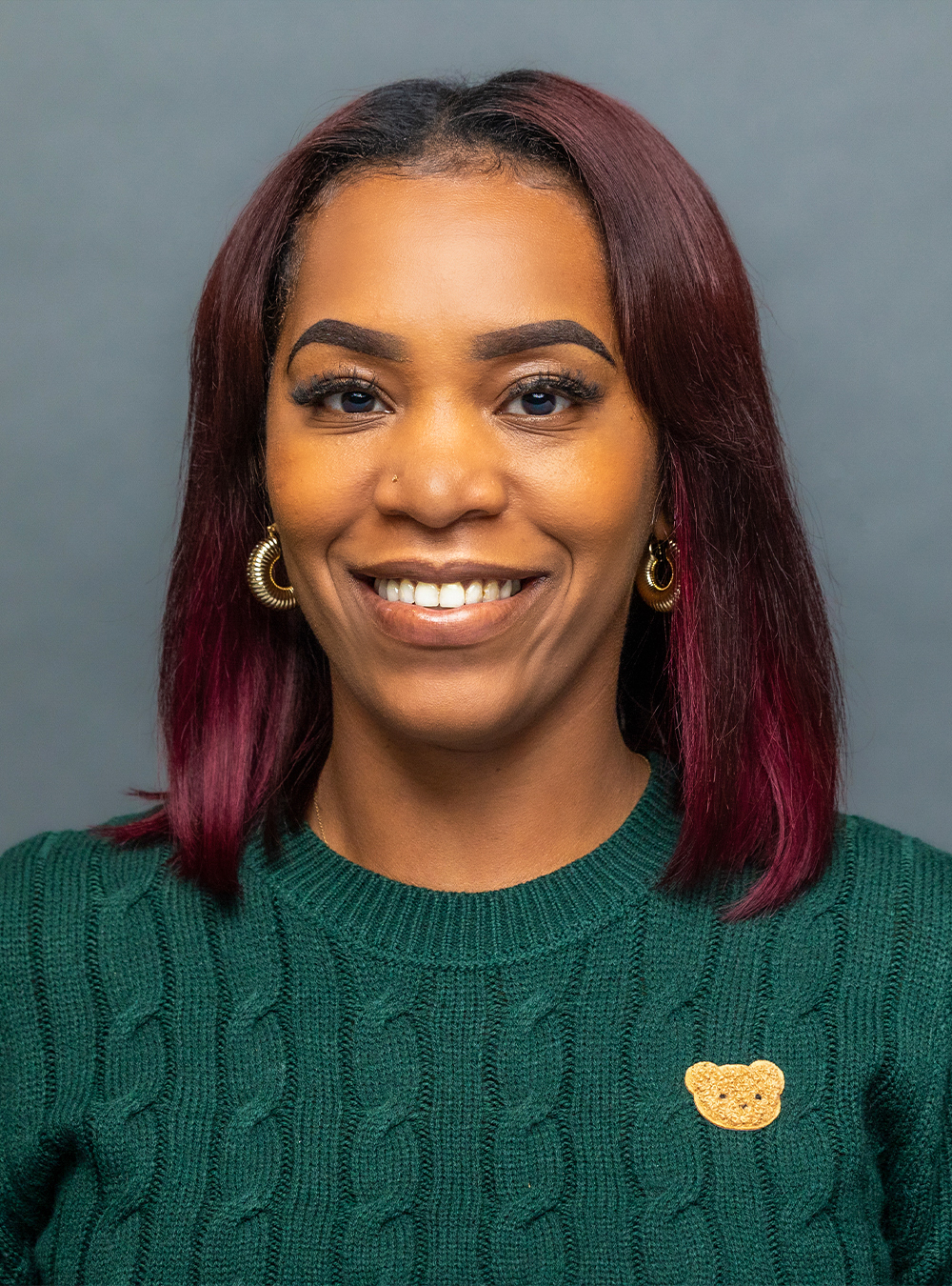Portrait of a smiling woman with shoulder-length burgundy hair wearing a green sweater against a neutral background