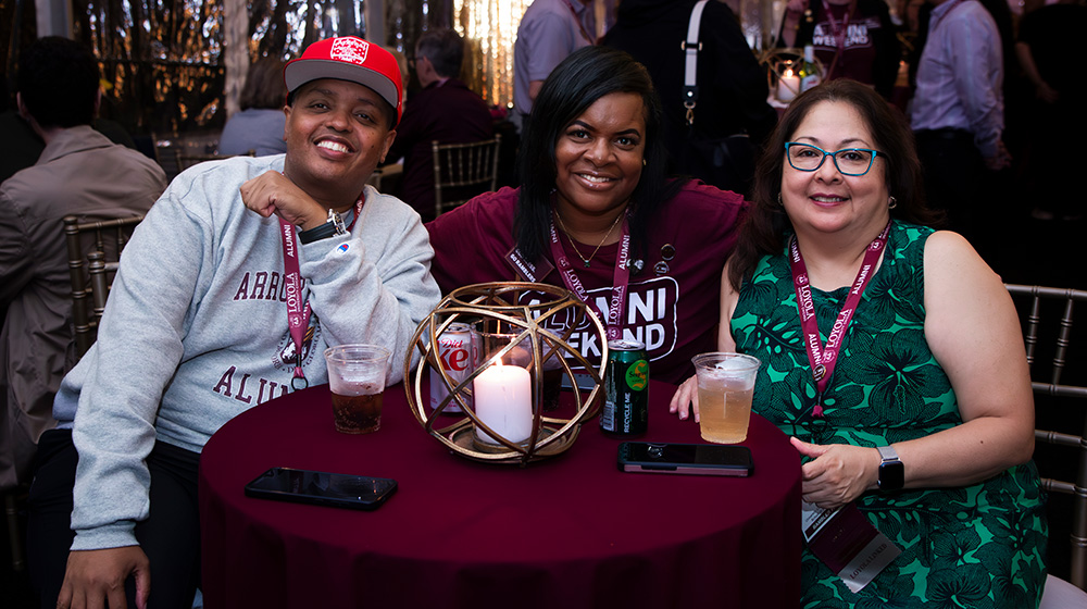 Three alumni in Loyola shirts enjoy drinks and conversation at an Alumni Weekend event hosted by an alumni affinity network.