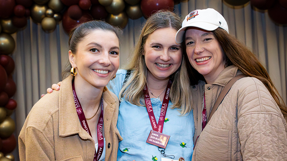 Three alumni in Loyola gear pose in front of an arch made of maroon and gold balloons at an Alumni Weekend event.
