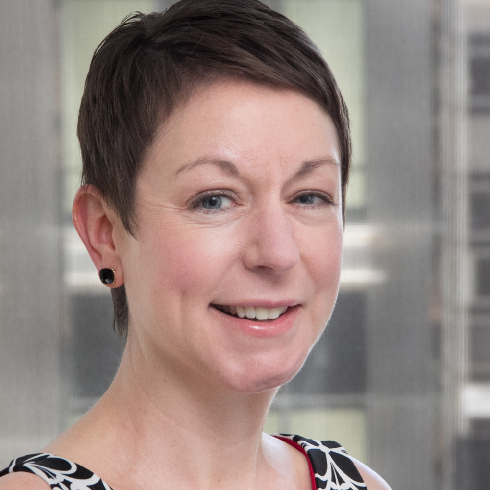 Professional headshot of a smiling woman with short dark hair wearing a patterned top, against a softly blurred indoor background