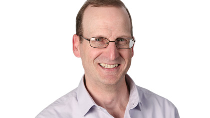 Smiling man with glasses and short brown hair wearing a light purple button-down.