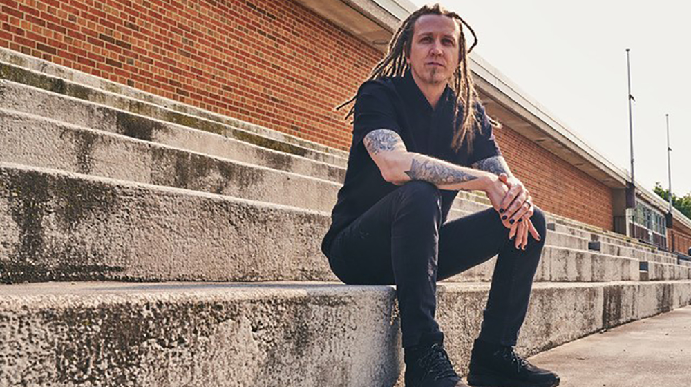 Man with long dreadlocks and tattoos sitting on outdoor concrete steps near a brick building.