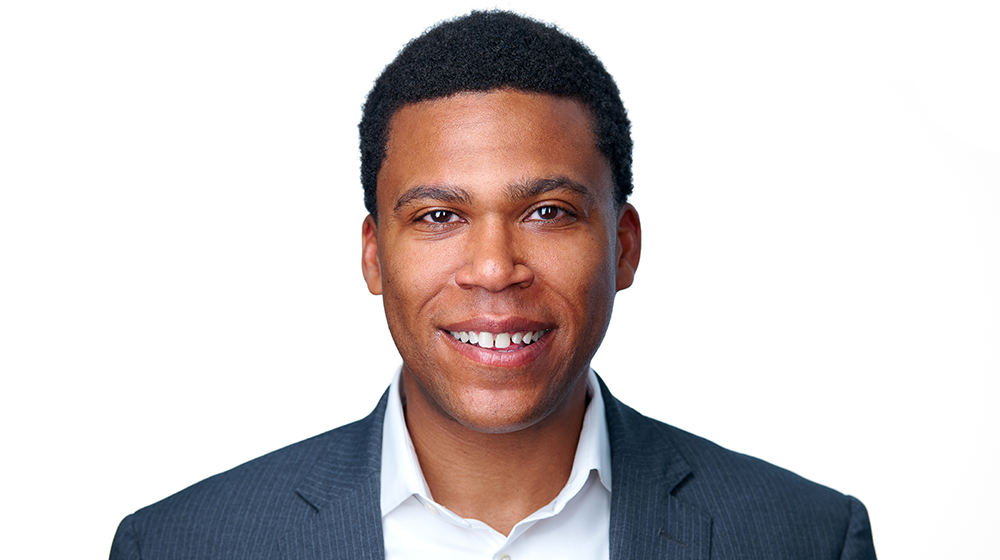 Portrait of a smiling man wearing a suit jacket and white shirt against a light background.
