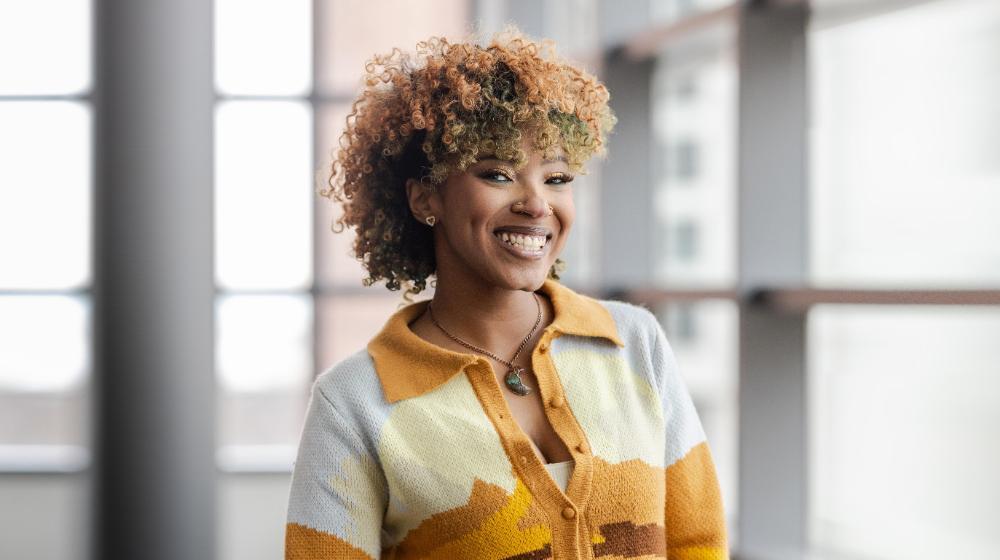 Arrupe College student with short curly hair standing indoors near large windows, wearing a patterned yellow and tan cardigan and looking confidently at the camera.