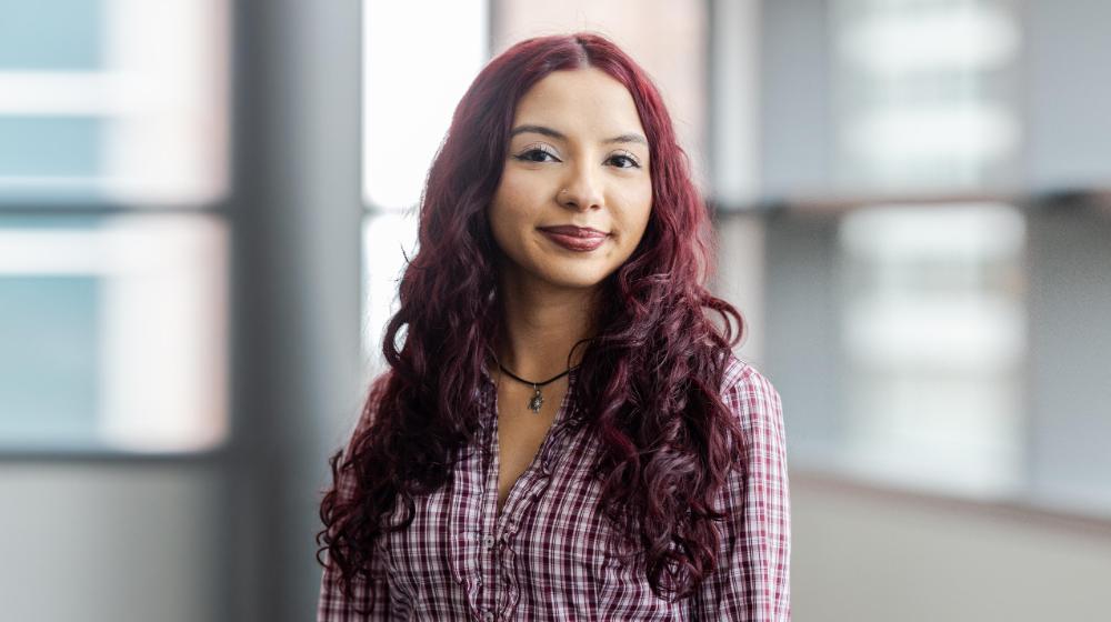 Arrupe College student with long burgundy hair standing indoors near large windows, wearing a plaid button-down shirt and looking calmly at the camera.
