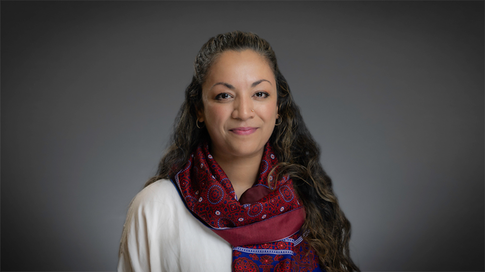 Portrait of a woman with long dark hair wearing a red patterned scarf against a gray background.