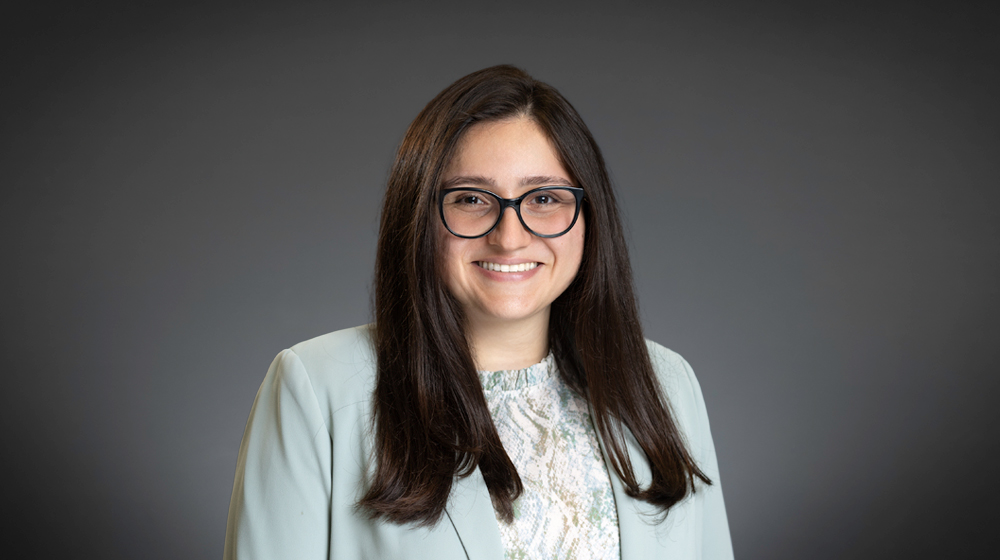 Smiling woman with long dark hair and glasses against a gray background.