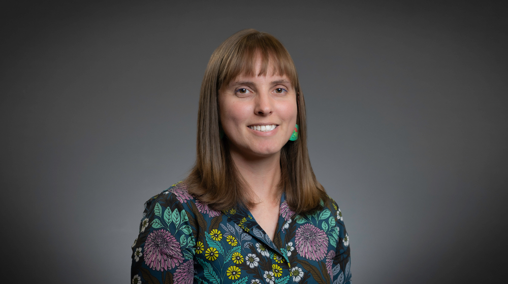 Portrait of a smiling woman with straight brown hair and bangs wearing a patterned blouse against a gray background.