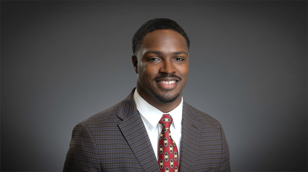 Portrait of a smiling man wearing a patterned suit jacket and tie against a gray background.