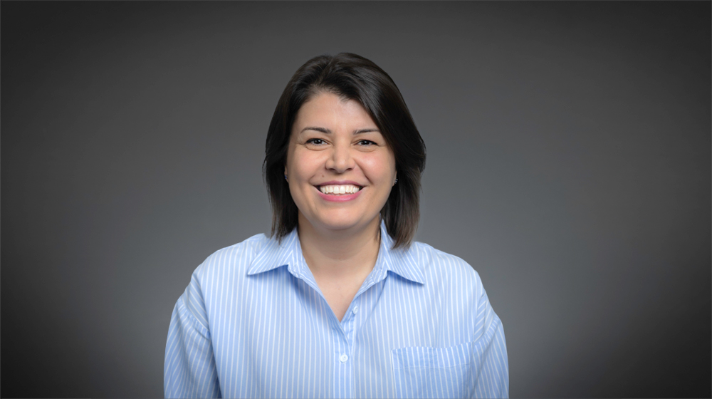 Smiling woman with short dark hair wearing a blue striped shirt against a gray background.