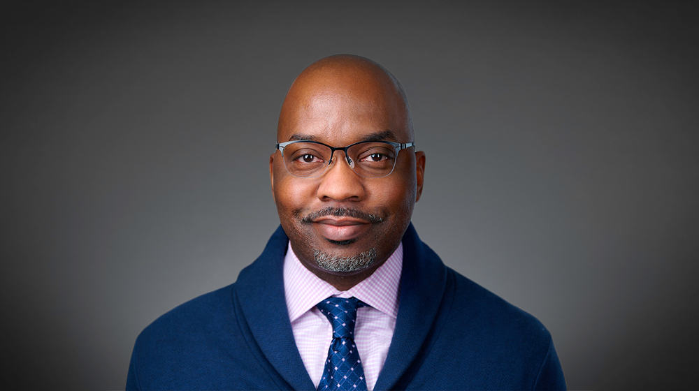 Man wearing glasses, tie, and sweater against a gray background.