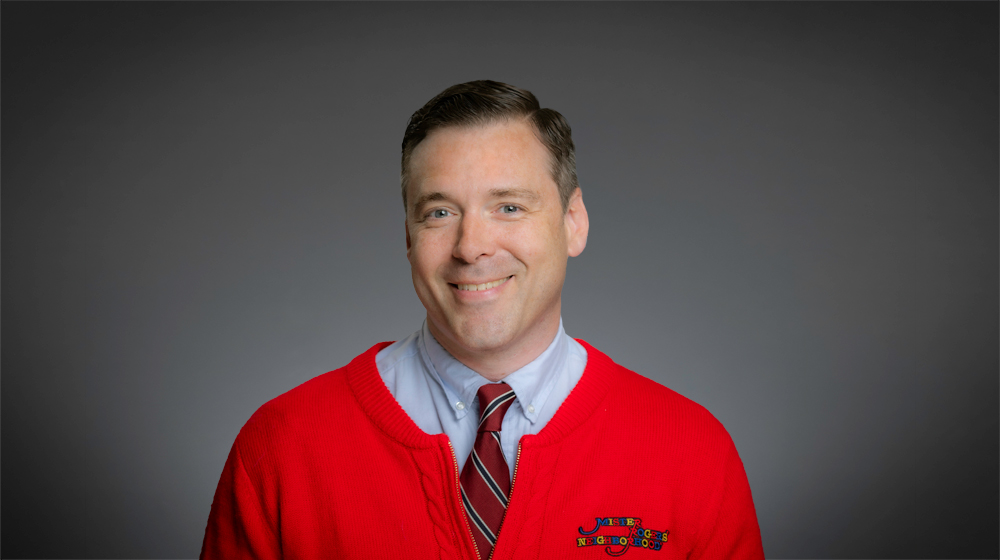 Smiling man wearing a red sweater and tie against a gray background.