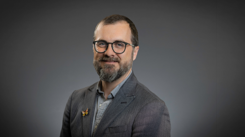 Portrait of a smiling man with short hair, beard, and glasses wearing a gray blazer against a gray background.