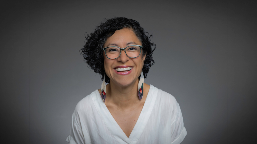 Portrait of a smiling woman with short curly hair and glasses wearing a white top against a gray background.