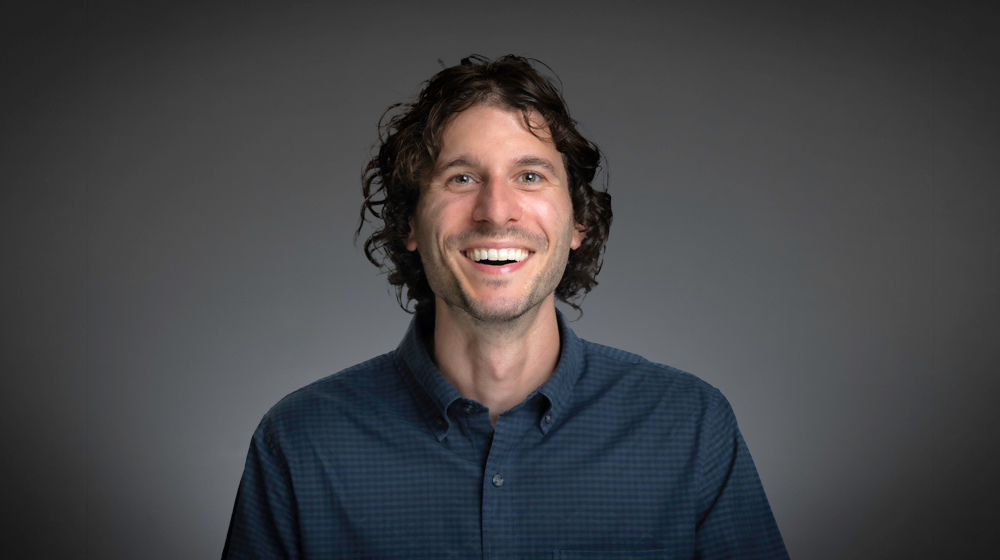 Smiling man with curly hair in a dark shirt against a gray background.