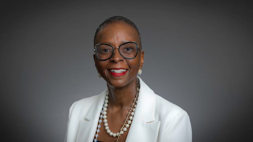 Smiling woman with short hair and glasses wearing a white blazer against a gray background.