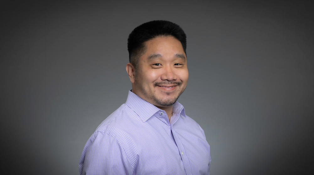 Smiling man in a light purple shirt against a gray background.