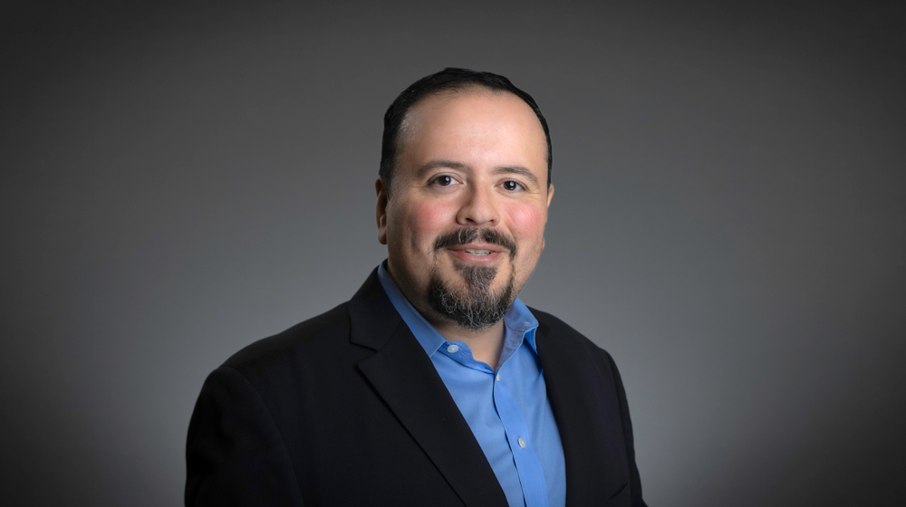 Man with a goatee in a blue shirt and blazer against a gray background.