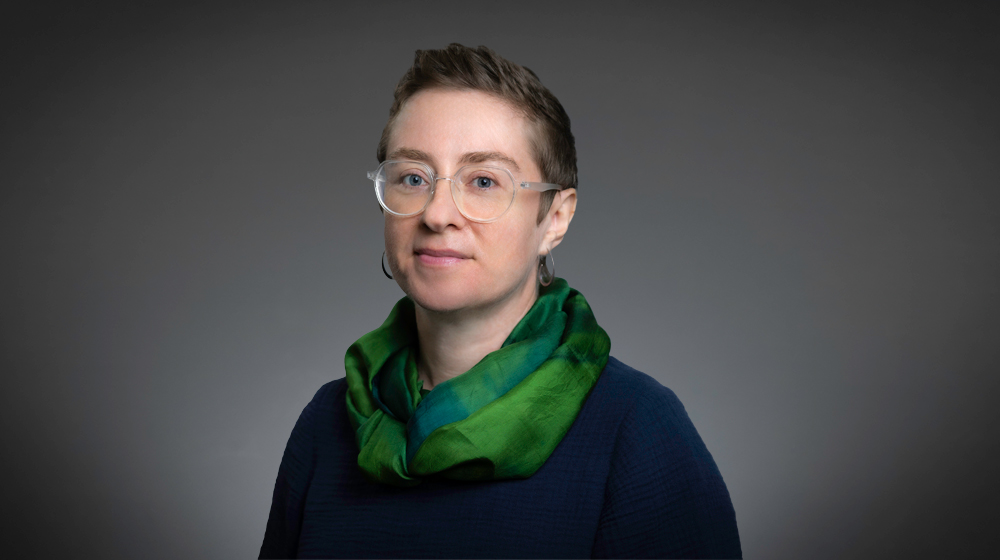 Person with short hair and glasses wearing a green scarf against a gray background.