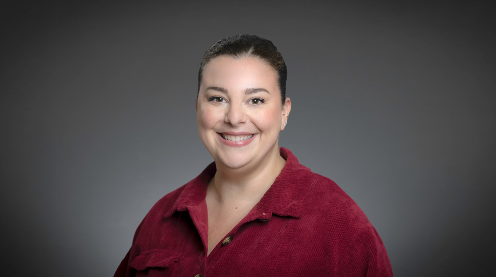 Portrait of a smiling woman with dark hair pulled back, wearing a red top, against a gray background.