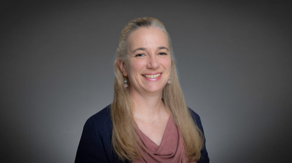 Portrait of a smiling woman with long blonde hair wearing a pink top and dark cardigan against a gray background.