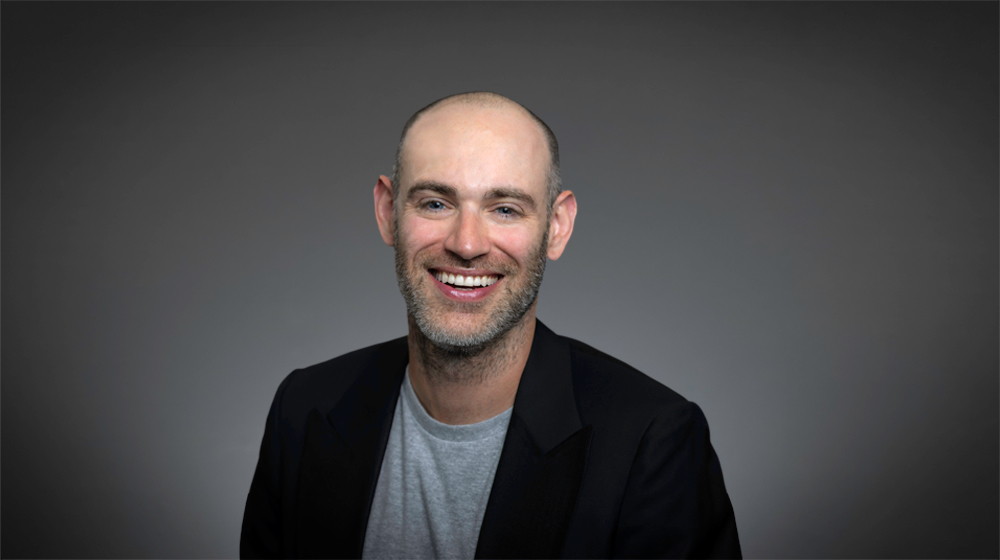 Smiling bald man in a blazer and gray shirt against a gray background.
