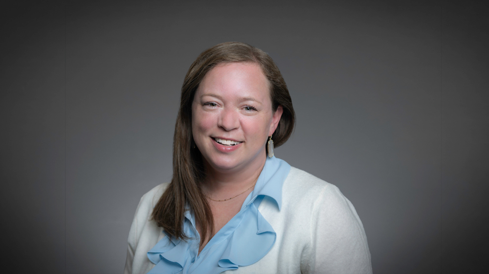 Portrait of a smiling woman with straight brown hair wearing a light blue blouse and white cardigan against a gray background.