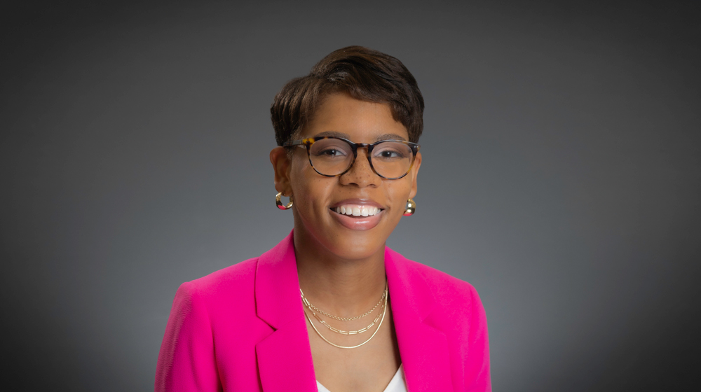 Portrait of a smiling woman with short hair and glasses wearing a bright pink blazer against a gray background.