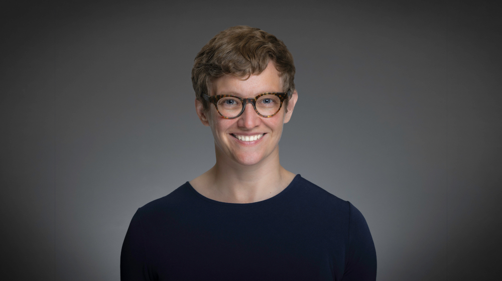 Smiling person with short hair and glasses against a gray background.