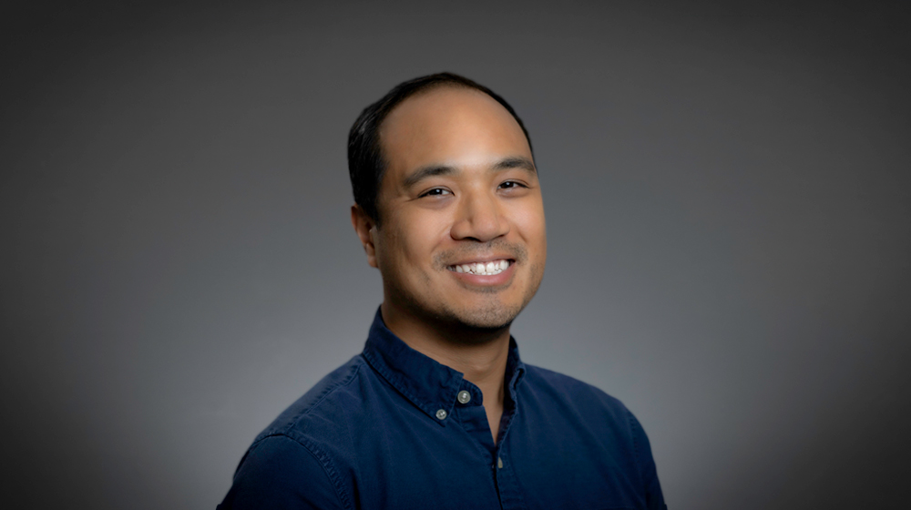 Portrait of a smiling man wearing a blue button-down shirt against a gray background.