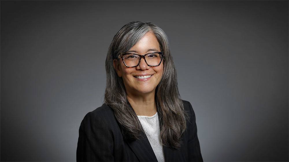 Portrait of a smiling woman with long gray hair and glasses wearing a dark blazer against a gray background.