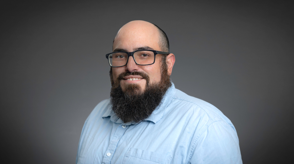 Man with glasses and a beard in a light blue shirt against a gray background.