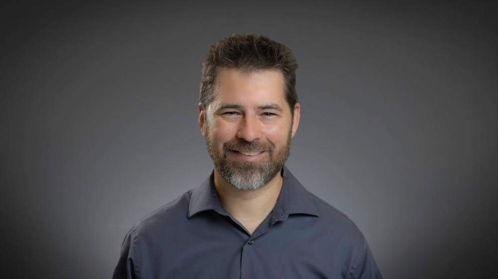 Portrait of a smiling man with short dark hair and a beard wearing a dark shirt against a gray background.