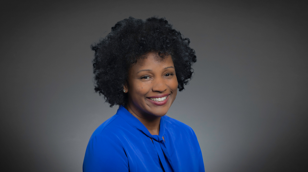 Smiling woman with curly hair in a blue blouse against a gray background.