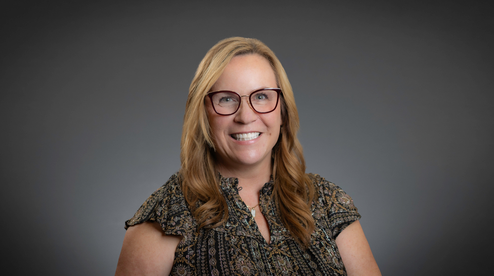 Portrait of a smiling woman with glasses and long blonde hair against a gray background.