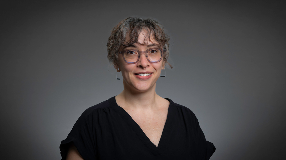 Portrait of a smiling person with short hair wearing a dark top against a dark background