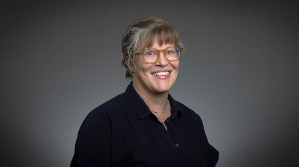 Portrait of a smiling woman with light brown hair and glasses wearing a dark top against a gray background.