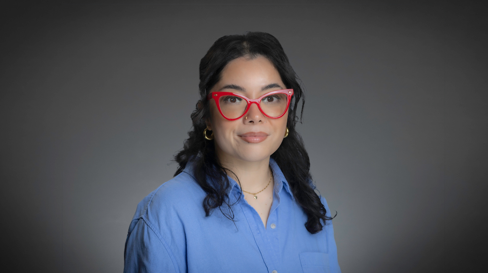 Person with dark hair wearing red glasses and a blue shirt against a gray background.