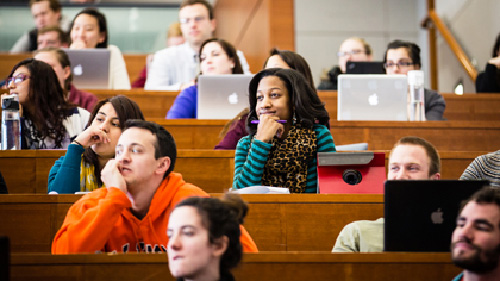  A group of students listen to an unseen lecturer. A young African American student wearing a green and black striped shirt sits in the center of the image with her hand to her chin.