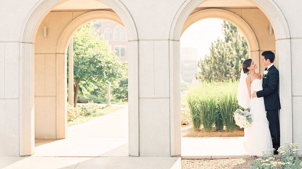 A bride and groom pose for a photo under the arches of the Madonna Della Strada Chapel on the Lake Shore Campus