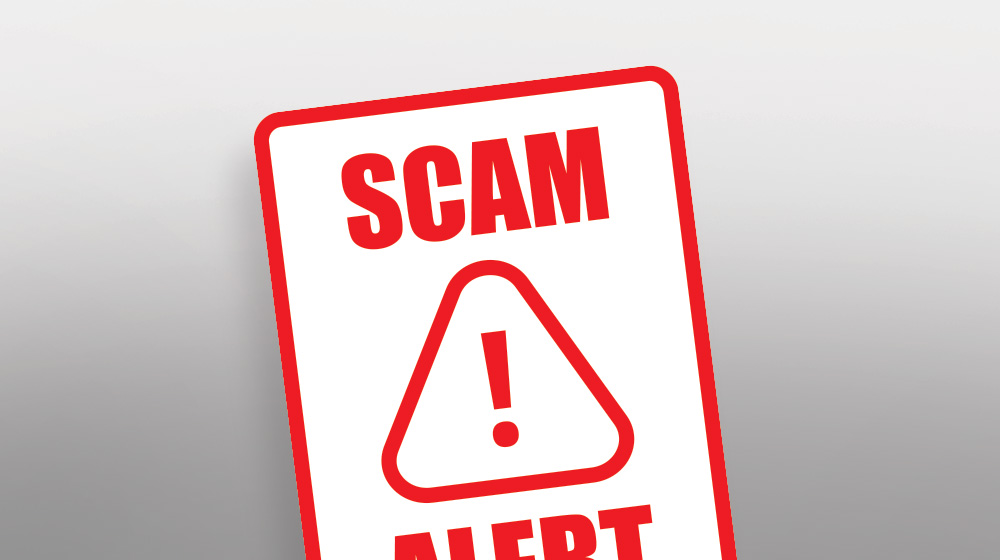 : A white sign with red block letters that read SCAM ALERT. A triangle with an exclamation sign in t