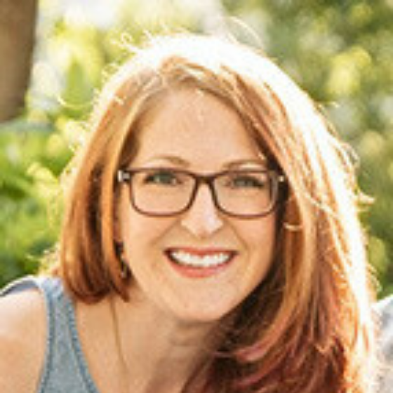 Smiling woman with long blonde hair and glasses outdoors, wearing a blue top, with blurred greenery in the background.