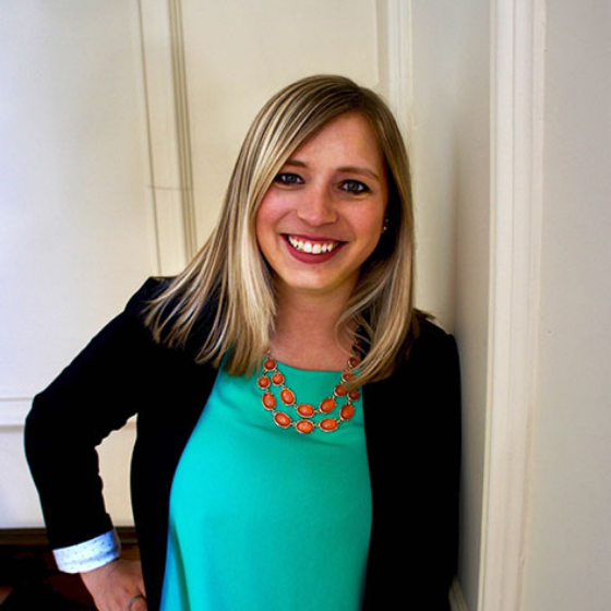 Smiling woman with straight blonde hair, wearing a teal top, black blazer, and an orange necklace, leaning against an off-white colored wall.