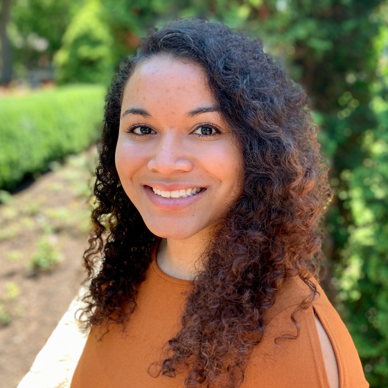 Smiling woman with curly, mid-length hair and an orange top standing outdoors, with greenery in the background.