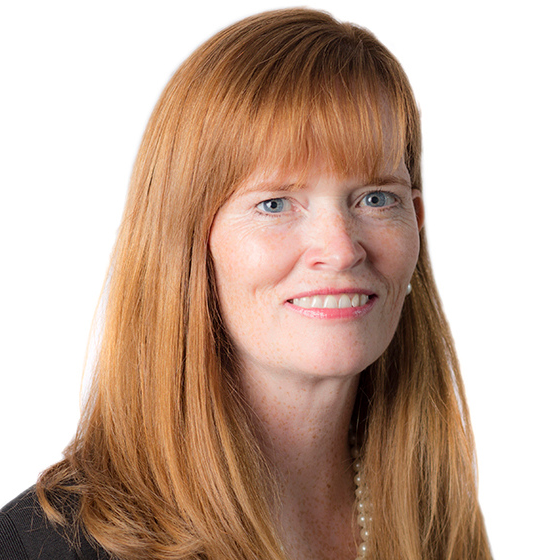 Smiling woman with long, red hair and bangs, wearing a black top and pearl necklace, against a solid white background.